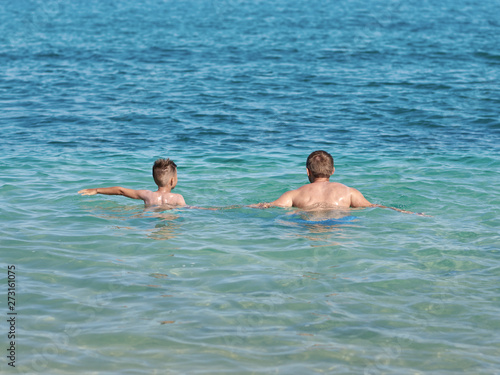 Boy is splashing in the sea with his dad. They are having fun during their holidays on the seacoast. Back view.