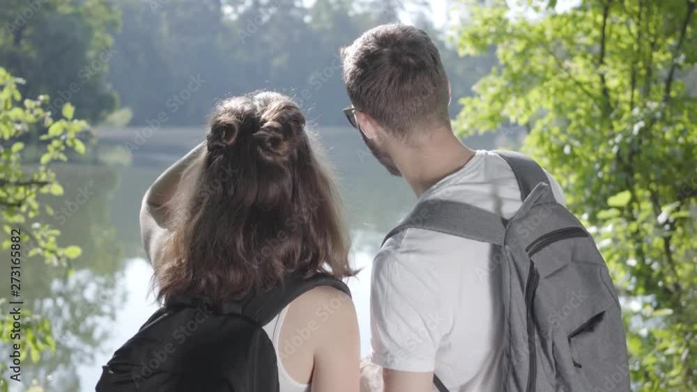 Portrait of guy and young cute girl standing on the riverbank in the forest with backpacks pointing away. The young man and woman hiking. Concept of camping. Leisure couples. Journey to nature. Portrait of guy and young cute girl standing on the riverbank in the forest with backpacks pointing away. The young man and woman hiking. Concept of camping. Leisure couples. Journey to nature.