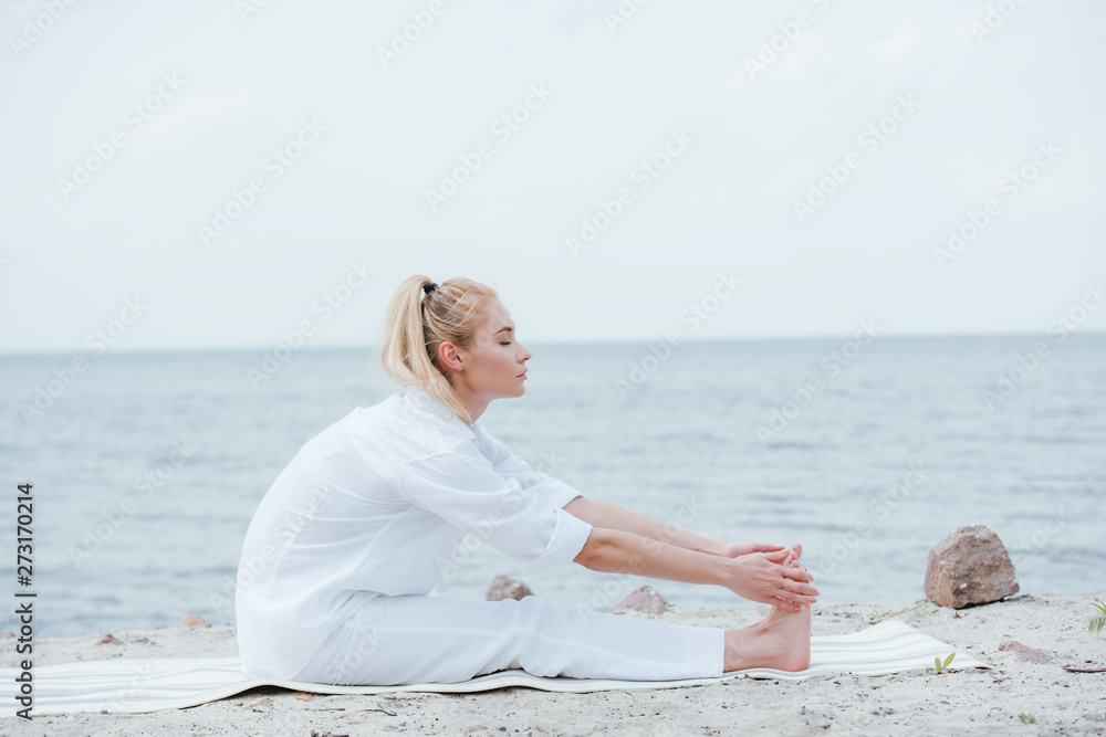 side view of attractive woman stretching while sitting on yoga mat near sea side view of attractive woman stretching while sitting on yoga mat near sea