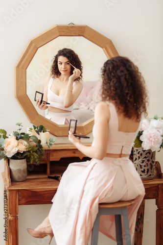 Young happy girl with curly hair does makeup in front of a vintage mirror. A beautiful woman in a pink dress paints her eyes.