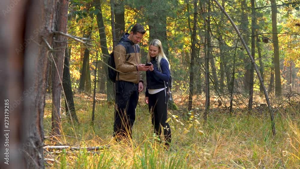 A hiking couple stands in the middle of a meadow in a forest on a sunny day and looks at a smartphone A hiking couple stands in the middle of a meadow in a forest on a sunny day and looks at a smartphone