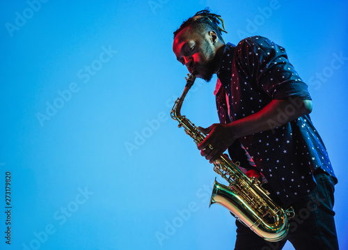 Fototapeta Young african-american jazz musician playing the saxophone on blue studio background in trendy neon light