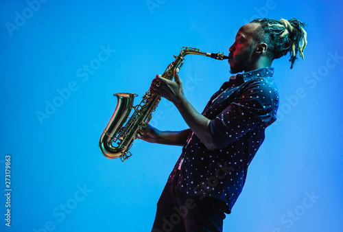 Photography Young african-american jazz musician playing the saxophone on blue studio background in trendy neon light