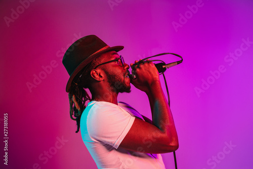Canvas Print Young african-american jazz musician with microphone singing a song on purple studio background in trendy neon light