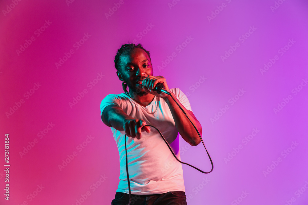 Young african-american jazz musician with microphone singing a song on purple studio background in trendy neon light. Concept of music, hobby, inspirness. Colorful portrait of joyful attractive artist
