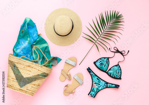 Bikini swimsuit with tropical print, silver glitter flat sandans, straw hat, wicker beach bag, sarong, tropical palm leaves on pink background. Overhead view of woman's swimwear and beach accessories.