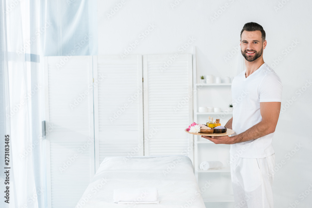 cheerful bearded man holding tray with bottles of oil, bowls with sea salt and flowers in spa center cheerful bearded man holding tray with bottles of oil, bowls with sea salt and flowers in spa center