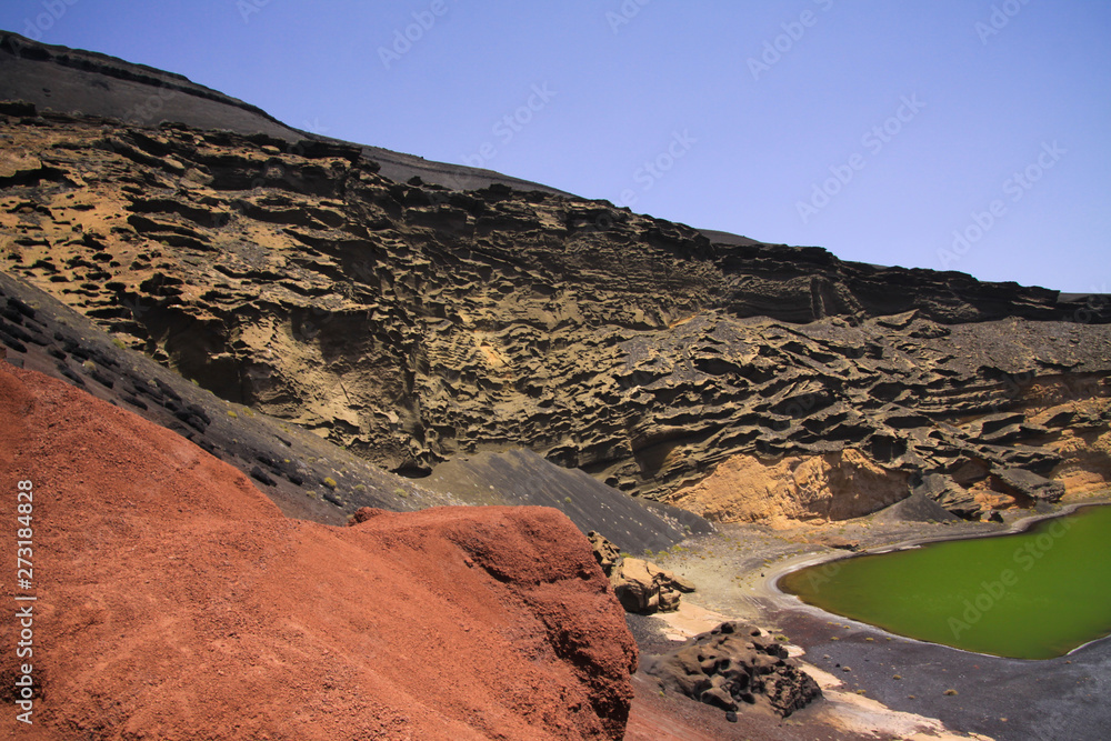 Fototapeta premium View over red rock on secluded deep green lake (Lago verde) in lagoon with black lava sand beach surrounded by impressive cliffs - El Golfo, Lanzarote