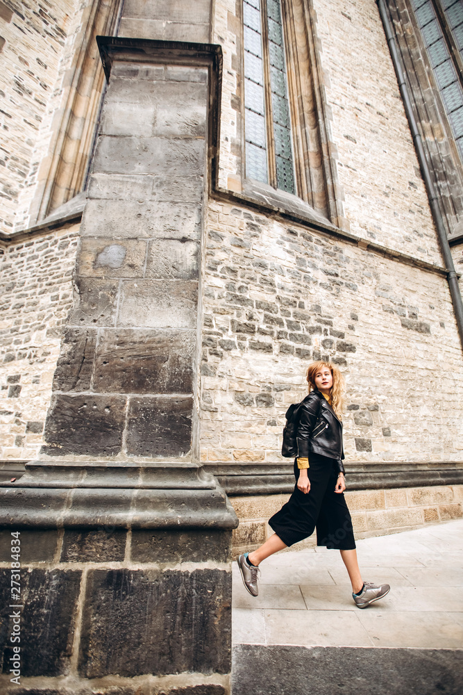 Young blonde with a backpack walking along a narrow cobbled street of Prague, Czech Republic. Stylish blonde girl in a leather jacket. Young blonde with a backpack walking along a narrow cobbled street of Prague, Czech Republic. Stylish blonde girl in a leather jacket.