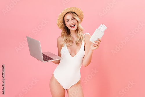 Happy excited young woman posing isolated over pink wall background dressed in swimwear beach concept using laptop computer holding passport with tickets.