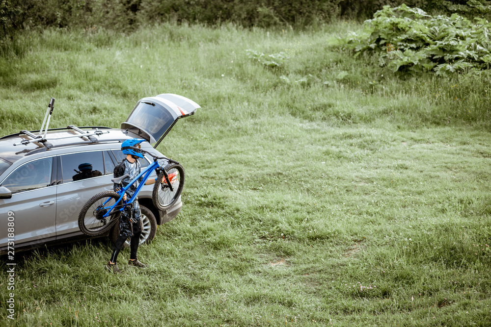 Fototapeta premium Profesjonalny rowerzysta wyciągający rower z bagażnika samochodu stojąc na zielonej łące w górach. Widok z góry