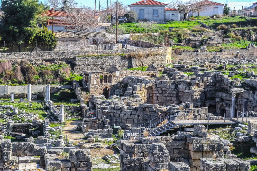 Excavated archaeological ruins in Ancient Corinth Greece with stairs and walkway for tourists and red tile roofed village houses behind