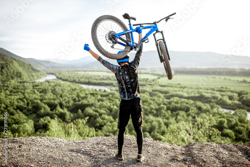 Professional well-equipped cyclist raising hands with bicycle while standing on the top of the mountain