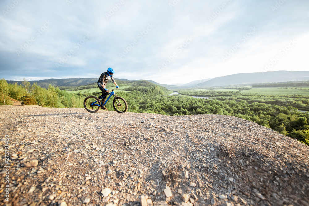 Fototapeta premium Professional well-equipped cyclist riding bicycle on the rocky mountains with beautiful landscape view during the sunset. wide view with copy space