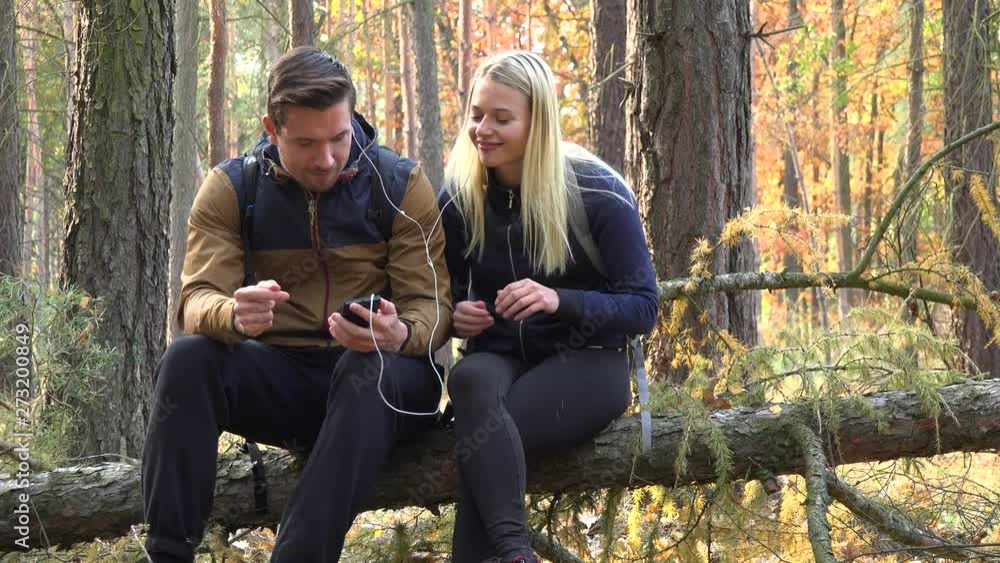 A hiking couple sits on a broken tree in a forest and listens to music on a smartphone A hiking couple sits on a broken tree in a forest and listens to music on a smartphone