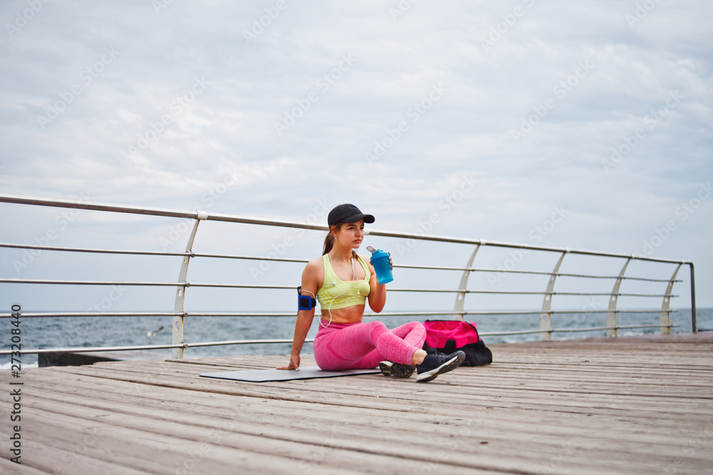 Young fit woman in sportwear sitting on a mat and drinking water on the beach