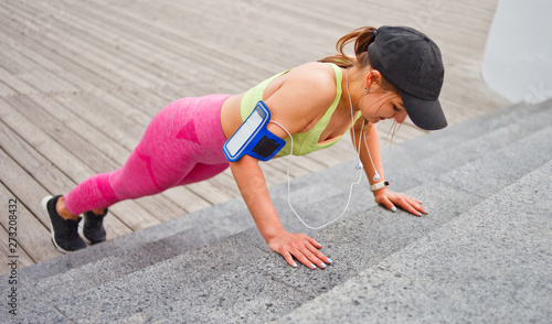 Cheerful fit woman in sportswear and cap doing push-ups from the stairs outdoors. Functional street workout.