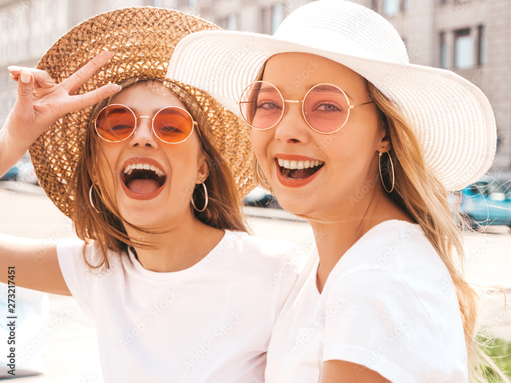 Portrait of two young beautiful blond smiling hipster girls in trendy summer white t-shirt clothes. Sexy carefree women posing on street background. Positive models having fun in sunglasses and hat