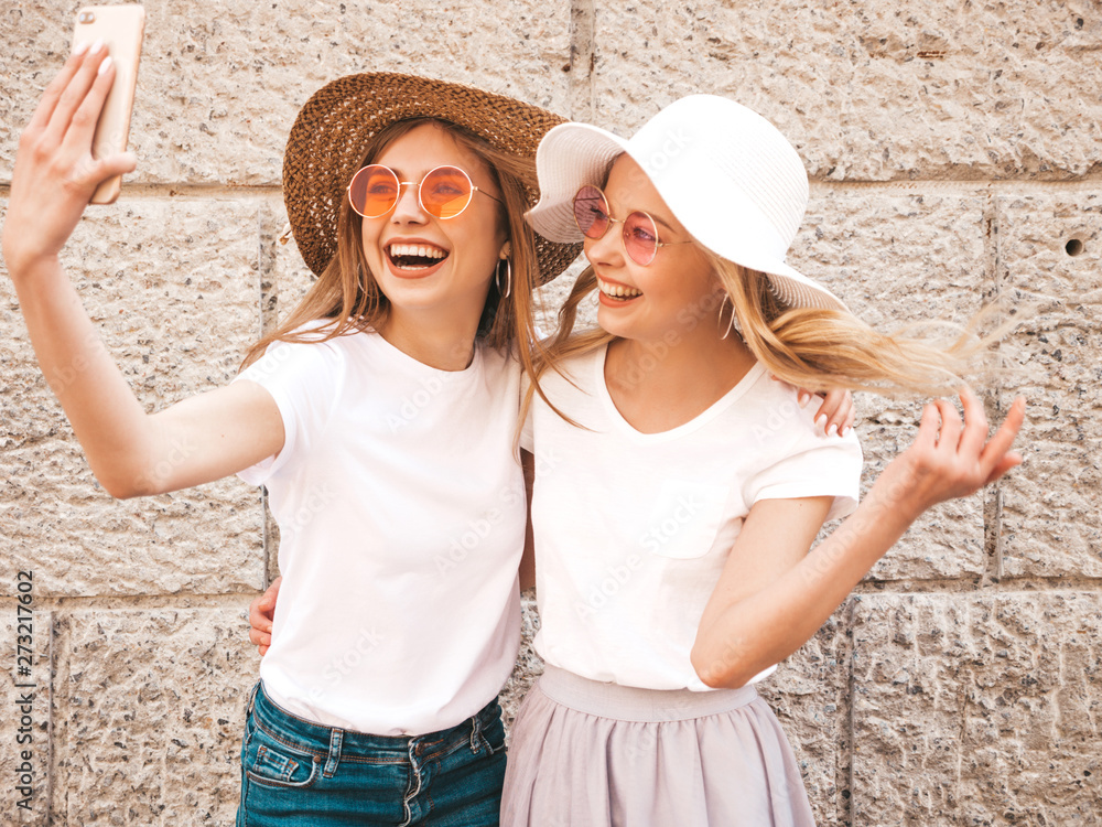 Two young smiling hipster blond women in summer white t-shirt. Girls taking selfie self portrait photos on smartphone.Models posing on street background.Female shows positive emotions