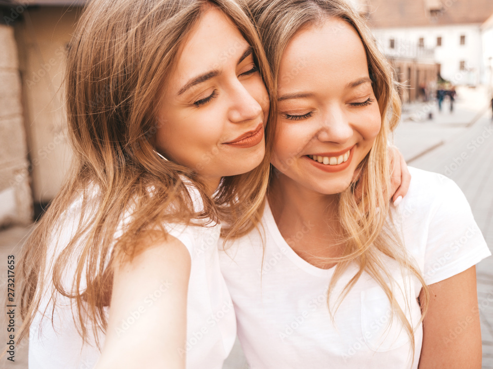 Portrait of two young beautiful blond smiling hipster girls in trendy summer white t-shirt clothes. Sexy carefree women posing on street background. Positive models having fun in sunglasses.Hugging Portrait of two young beautiful blond smiling hipster girls in trendy summer white t-shirt clothes. Sexy carefree women posing on street background. Positive models having fun in sunglasses.Hugging