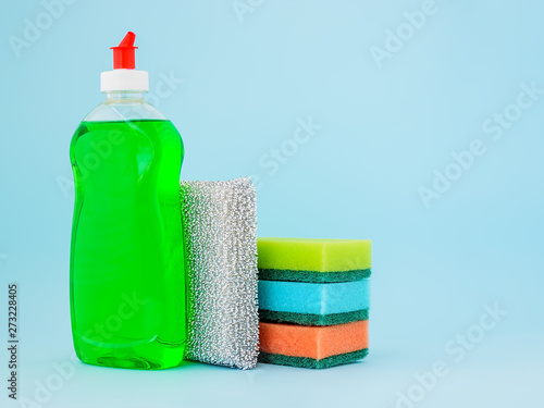 Fotografie Transparent bottle with green dishwashing liquid and four foam sponges of different colours on a blue background