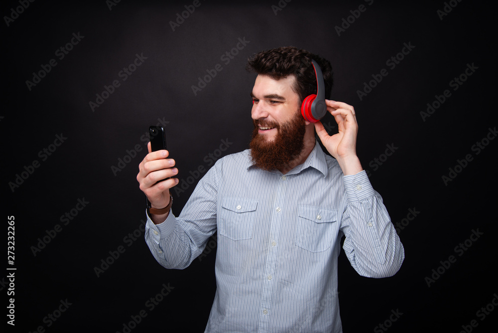Photo of handsome bearded man using a mobile phone and headphoneas , standing over dark isolated backgound Photo of handsome bearded man using a mobile phone and headphoneas , standing over dark isolated backgound