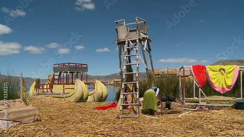 Medium low-angle still shot of Uros village compound with a ladder leaning on lookout tower, coloured traditional skirts, reed boat, and a woman in front of a  cooking stove, Uros Village, Peru