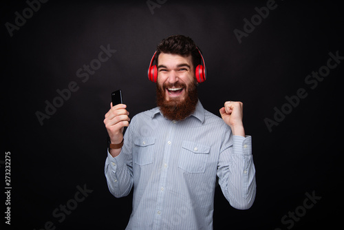 Photo of cheerful bearded young man, celebrating with arms up, while using a mobile phone and headphoneas