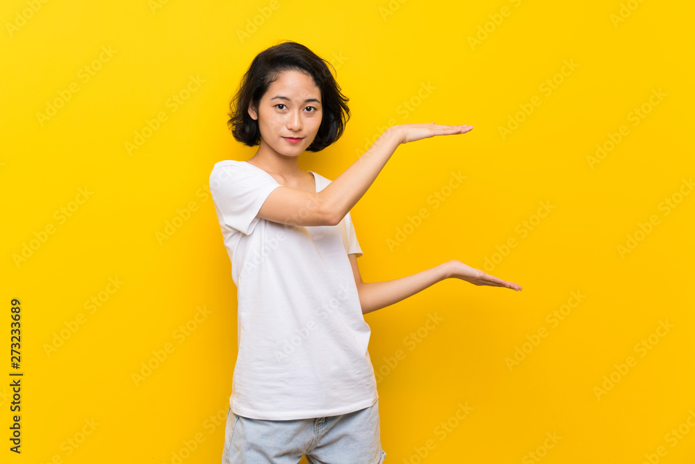 Asian young woman over isolated yellow wall holding copyspace to insert an ad Asian young woman over isolated yellow wall holding copyspace to insert an ad