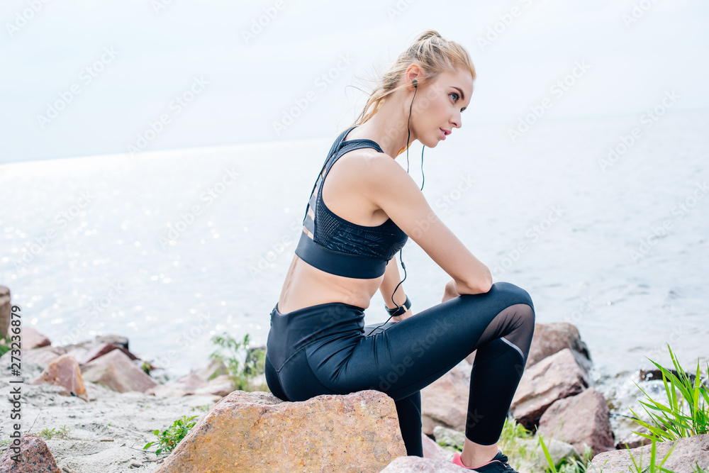 athletic young woman sitting on stone and listening music in earphones near sea