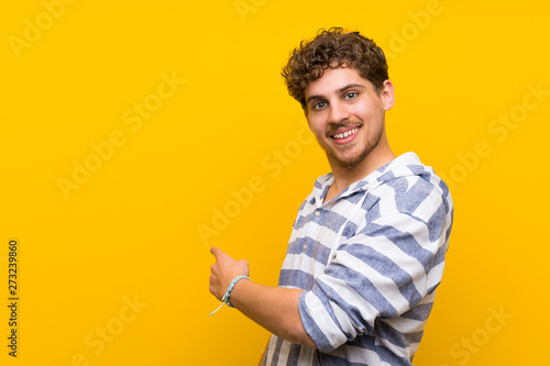 Blonde man over yellow wall pointing back with the index finger