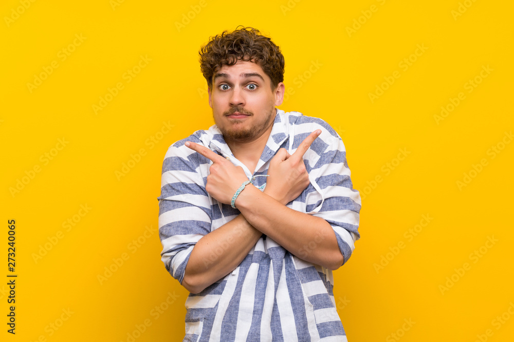 Blonde man over yellow wall pointing to the laterals having doubts Blonde man over yellow wall pointing to the laterals having doubts