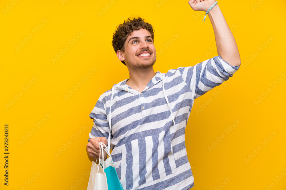 Blonde man over yellow wall holding a lot of shopping bags in victory position Blonde man over yellow wall holding a lot of shopping bags in victory position