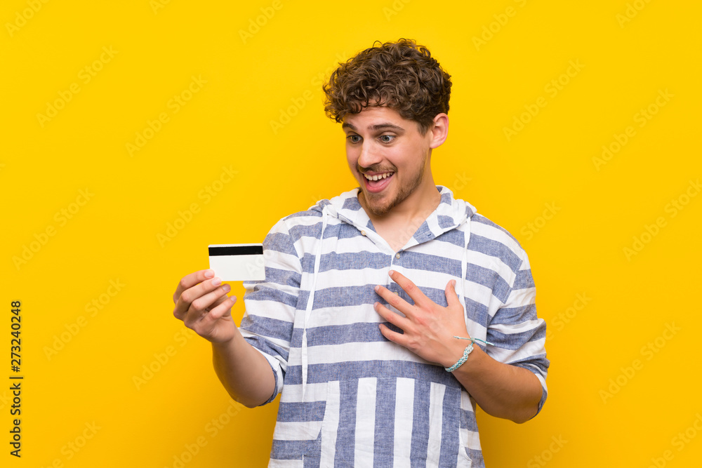 Blonde man over yellow wall holding a credit card and surprised Blonde man over yellow wall holding a credit card and surprised