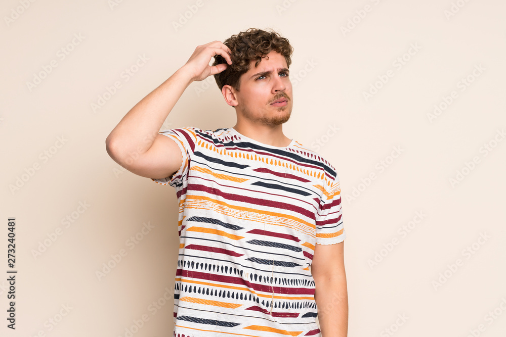 Blonde man over isolated wall having doubts while scratching head Blonde man over isolated wall having doubts while scratching head