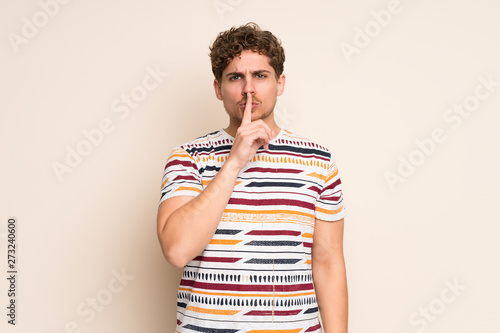 Blonde man over isolated wall showing a sign of silence gesture putting finger in mouth
