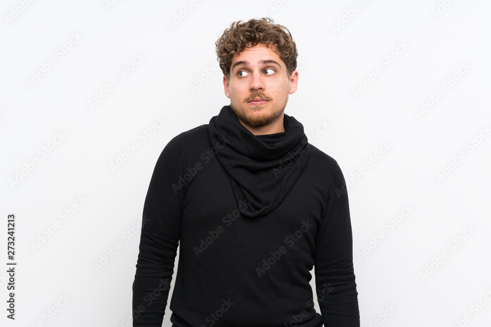 Blonde man over isolated white wall making doubts gesture looking side Blonde man over isolated white wall making doubts gesture looking side