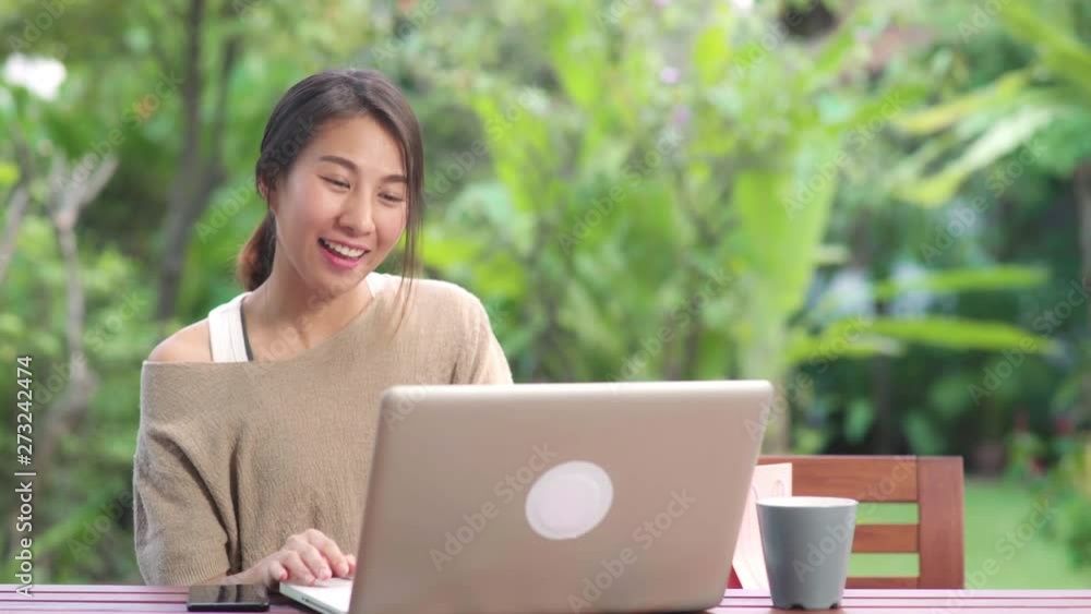 Asian woman using laptop video conference with friends, female relax feeling happy showing shopping bags sitting on table in the garden in morning. Lifestyle women relax at home concept. Asian woman using laptop video conference with friends, female relax feeling happy showing shopping bags sitting on table in the garden in morning. Lifestyle women relax at home concept.
