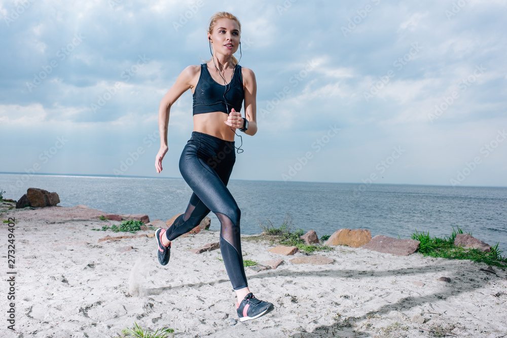 sportive blonde woman running and listening music in earphones near sea