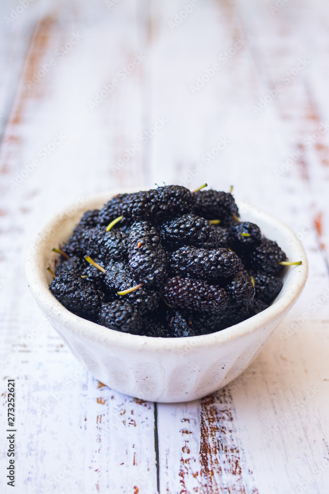 Sweet ripe black mulberry berries in a bowl. On wooden white table. Sweet ripe black mulberry berries in a bowl. On wooden white table.