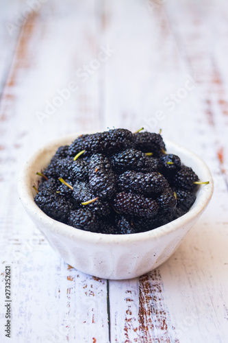 Sweet ripe black mulberry berries in a bowl. On wooden white table.