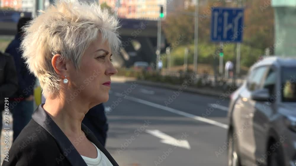 A middle-aged woman stands by a road in an urban area and looks around - closeup A middle-aged woman stands by a road in an urban area and looks around - closeup