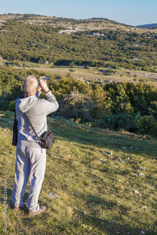 A man with blond hair stands on Mount Ai-Petri in the Crimea against the background of a forest and a plateau and looks through a telescope