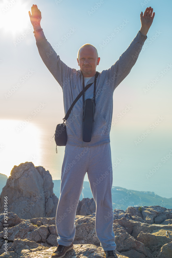 a man with blond hair and arms raised up, a telescope hangs on his neck, stands on the edge of Ai-Petri mountain in the Crimea against the background of the sea and rocks at sunrise a man with blond hair and arms raised up, a telescope hangs on his neck, stands on the edge of Ai-Petri mountain in the Crimea against the background of the sea and rocks at sunrise