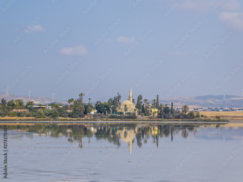 Hala Sultan Tekke seen from a distance. The mosque is surrounded by lush setting - palm trees and smaller bushes. Clear reflection of the mosque in the calm surface of a Larnaca Salt Lake.