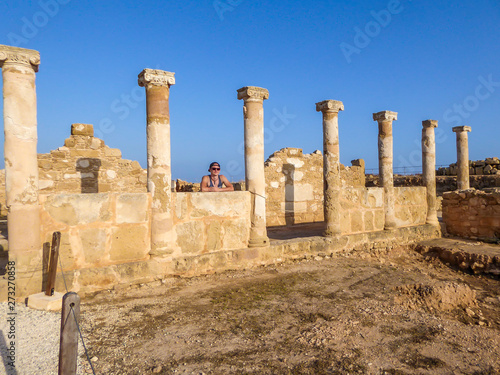 Young man standing between the ruins of an ancient temple, what is left are temple columns, Kato Paphos Archeological Park. Sandy color of the construction. Clear and blue sky. © Chris
