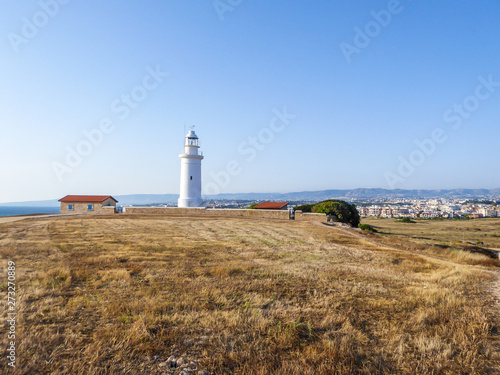 A white lighthouse located by the shore in Kato Paphos Archeological Park. Next to it are few small houses. The ground is covered with dried grass. In the back there is a big city agglomeration.