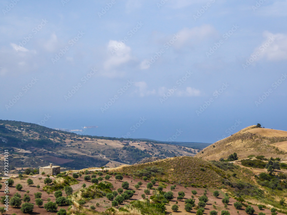 An idyllic view of a hilly fields in Cyprus. Slopes are overgrown with grains and small bushes. In the back some smaller mountains are visible. Great overcast, spreading all over the sky.