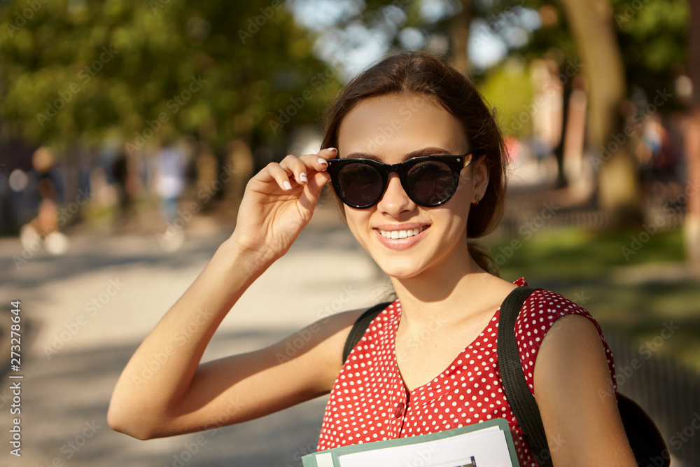 Youth, summertime, style and happiness concept. Cheerful fashionable pretty girl with slender body and cute happy smile holding hand on her black sunglasses, spending nice time outdoors, walking Youth, summertime, style and happiness concept. Cheerful fashionable pretty girl with slender body and cute happy smile holding hand on her black sunglasses, spending nice time outdoors, walking