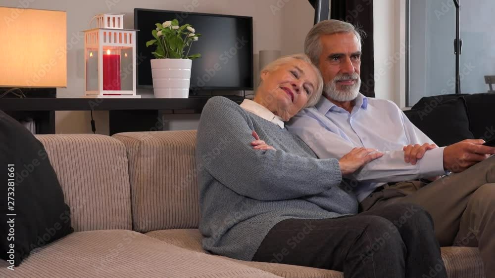 An elderly couple sits in a living room in an apartment and watches TV, which is off the camera An elderly couple sits in a living room in an apartment and watches TV, which is off the camera